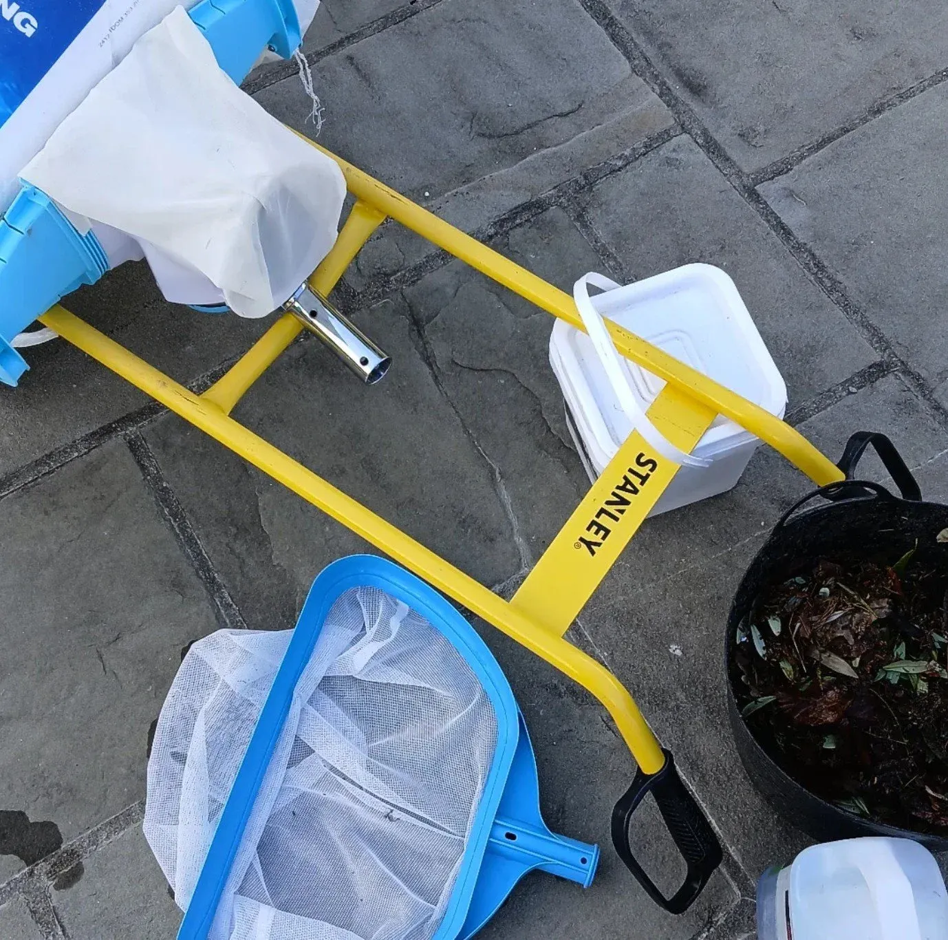 The valet kit on a paved courtyard: a yellow trolley, chemical bucket, leaf scoop and nets.